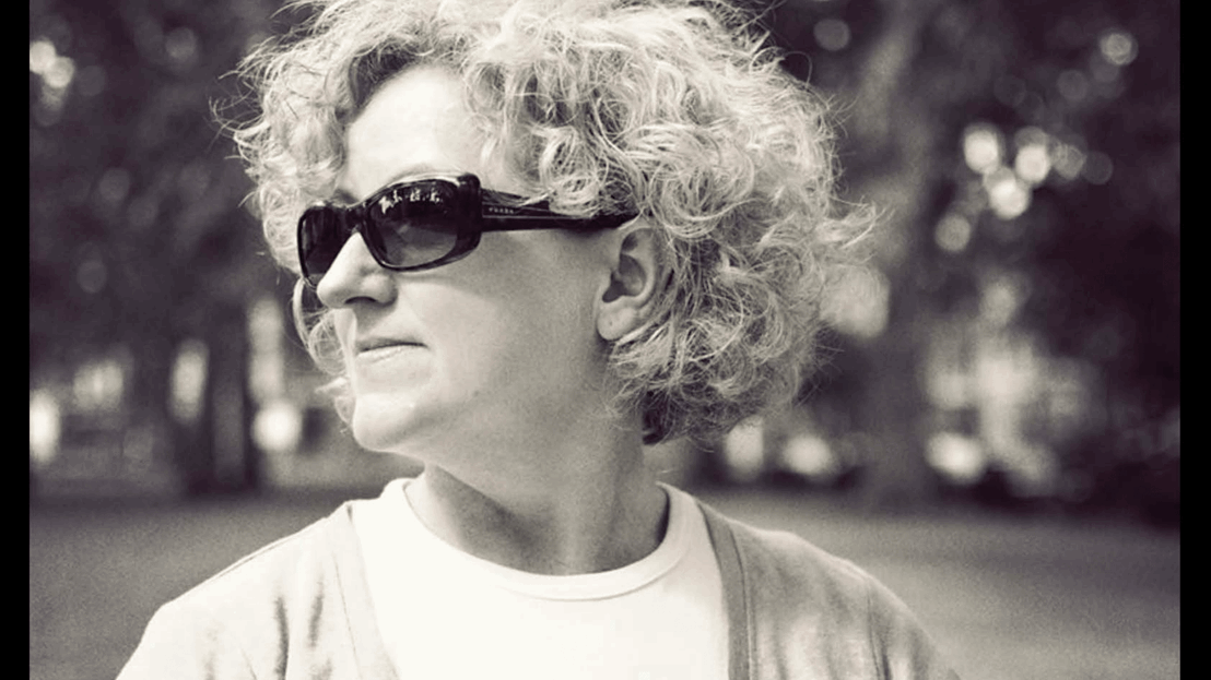 A monotone image of a person wearing sunglasses turned to the side with a blurry park and trees behind her.